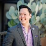 Portrait photo of man with lavender shirt and grey blazer. He has short hair and is smiling. 