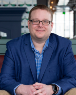 Portait photo of a smiling man with checkered blue shirt, royal blue blazer, glasses, and short hair.